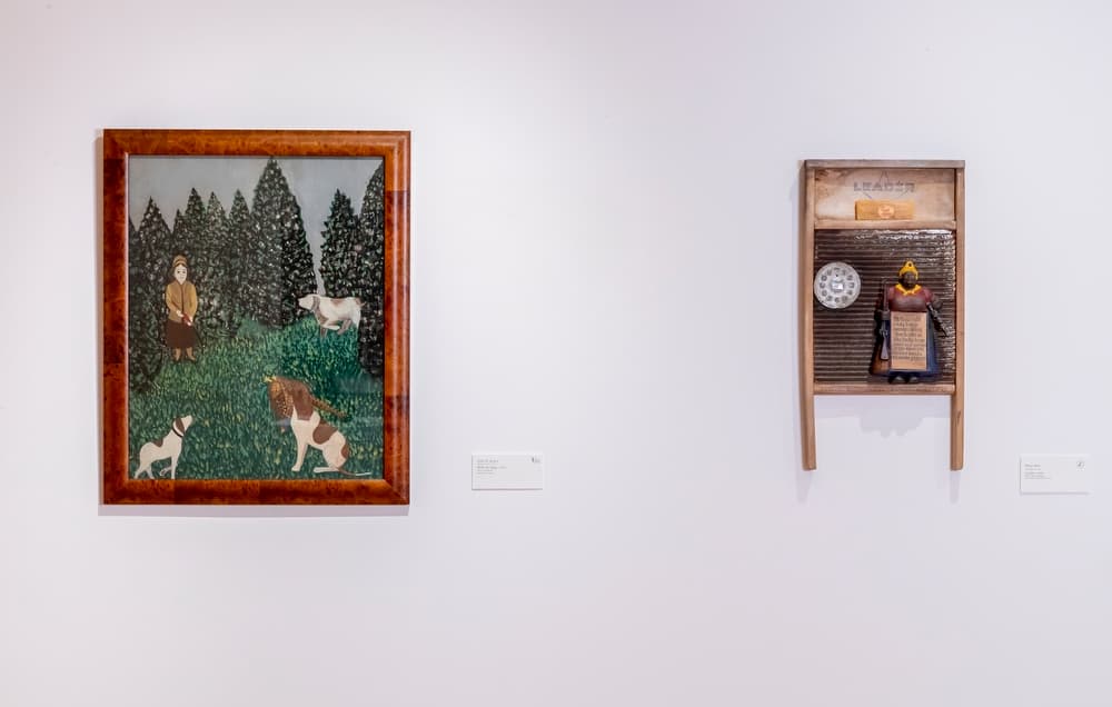 Two works of art hung side by side, one a framed painting of a women hunting with three dogs and the other, a multimedia work including a washboard and a figurine of a Black female.
