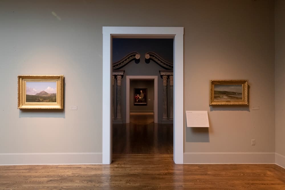 A series of interconnected galleries shown through doorways. Two landscape paintings flank one doorway and a portrait hangs in the background
