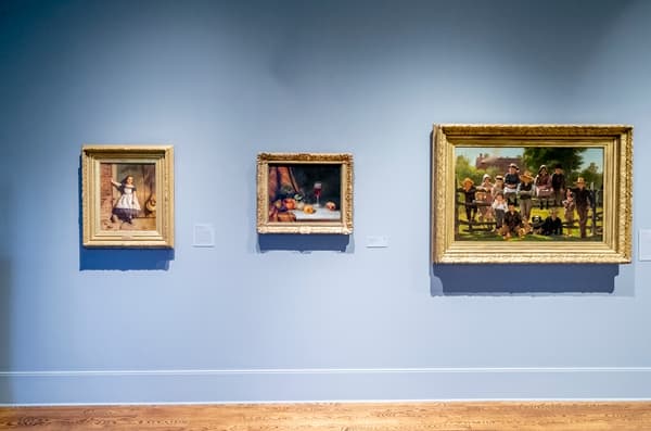 Three framed paintings hang side by side on a light blue gallery wall above a wooden floor.