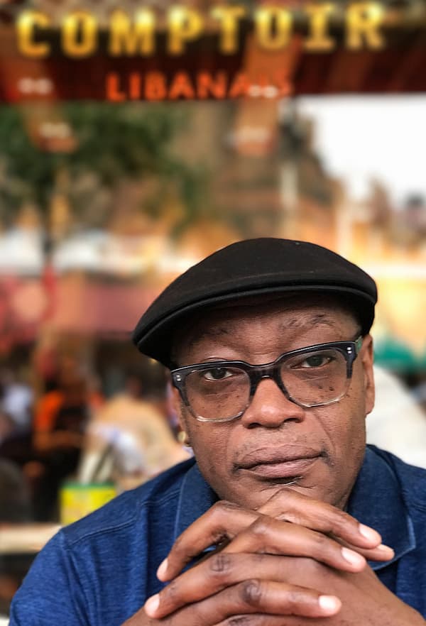 A man with glasses and a flat cap with a blurred street scene in the background.