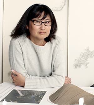 Maya Lin in her studio, wearing glasses in a light gray sweater leans on a table with a topographical model and photograph.