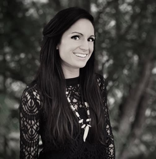 Black and white portrait of a person in a lace dress with a statement necklace, set against a blurred background of foliage.