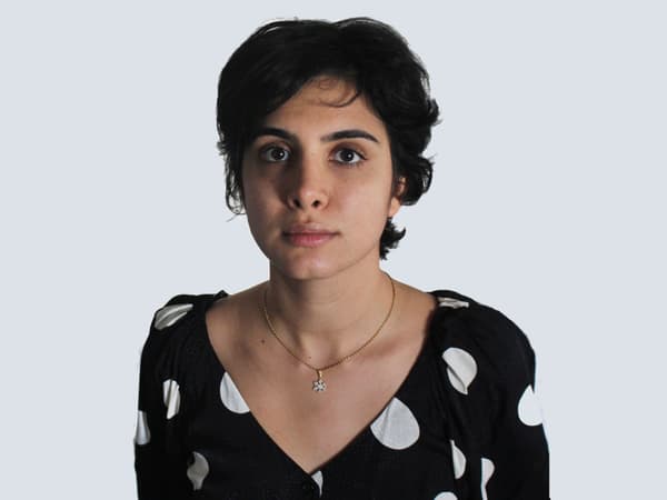 Person with short dark hair in a black, polka-dotted top and a gold necklace against a light background.
