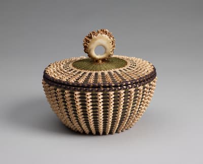 A circular woven basket with a patterned lid and a ring-shaped handle against a gray background.