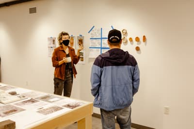 Two people in a room with ceramic art and documents displayed on the wall.