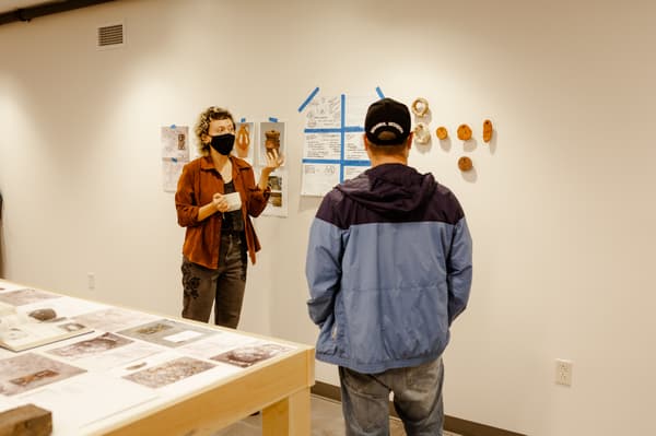 Two people in a room with ceramic art and documents displayed on the wall.
