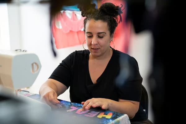 A person works at a sewing machine on a colorful fabric with "HEAL" in multicolored letters.
