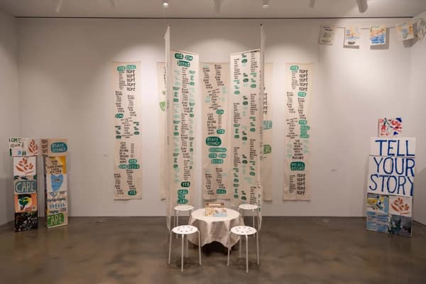 The image showcases an art installation in a gallery space. Five long vertical fabric banners are hanging from the ceiling, each featuring a mix of words in bold black and green lettering on a cream background. Below the banners, a small round table is surrounded by four white stools. The table is covered with a white tablecloth, and a small wooden box or container is placed on top. On the left and right sides of the room, there are additional art pieces, including some with leaf motifs and various painted texts. The right side features a large canvas with the words "TELL YOUR STORY" in blue letters. The gallery floor is polished concrete, and the walls are white, giving the room a clean, minimalist look.