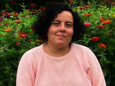 Photograph of Heather Flor Cron wearing a pink sweater in front of a lush green and floral background.