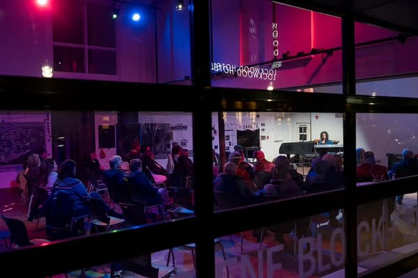 In a photo taken from outside through a window, a group of people seated in chairs watch a musician on stage. The room is colorful with blue and red lighting, and the musician is illuminated by a spotlight.