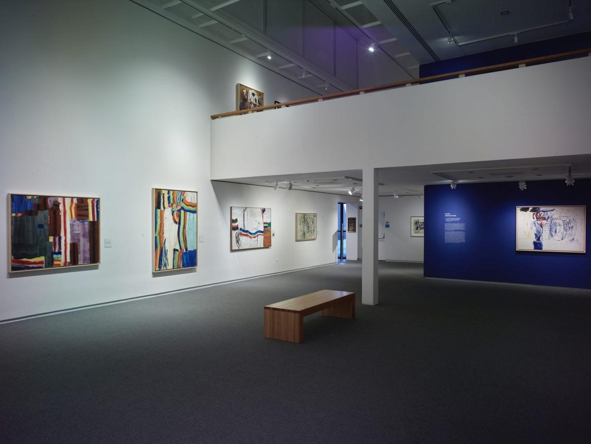 Art gallery interior with abstract and contemporary paintings on white and blue walls with a wooden bench in the center.