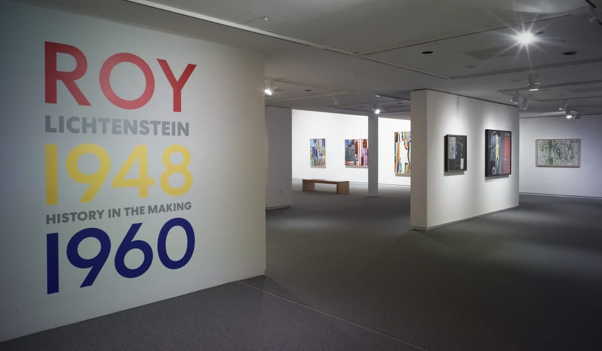 Art gallery interior with abstract and contemporary paintings on white walls, the front wall reading, "Roy Lichtenstein: History in the Making, 1948-1960."