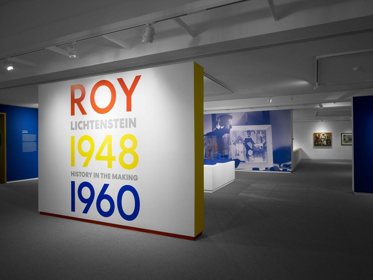 Art gallery interior with abstract and contemporary paintings on white walls, the front wall reading, "Roy Lichtenstein: History in the Making, 1948-1960."