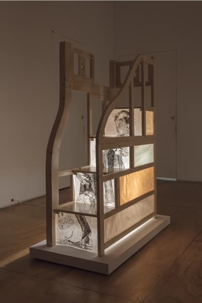 A wooden sculptural installation with illuminated translucent panels displaying drawings of human figures.