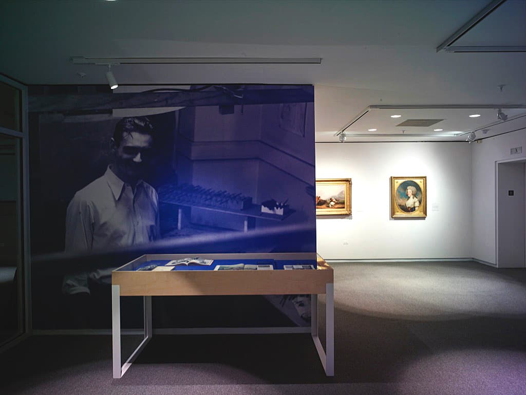 Art gallery interior with abstract and contemporary paintings on white and walls. The front blue wall is overlaid with an image of a man, and in front of it, is a plexiglass table displaying books.