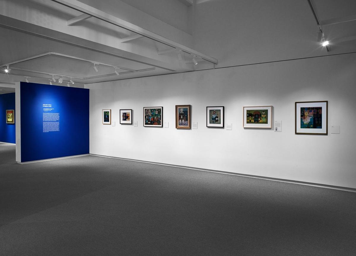 Art gallery interior with abstract and contemporary paintings on white walls next to a blue wall with overlaid wall text.