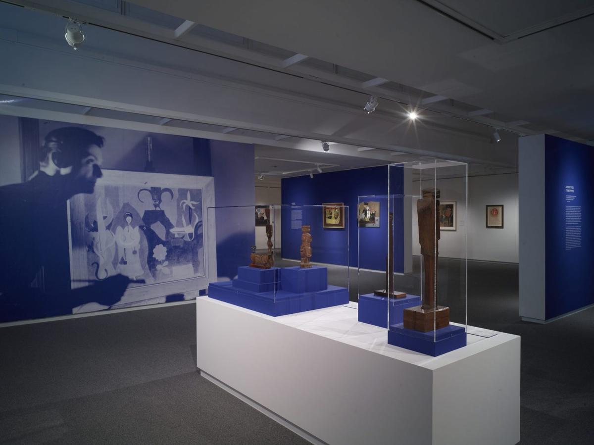 Art gallery interior with abstract and contemporary paintings on white and blue walls with a white table in the center with multiple objects in plexiglass boxes.
