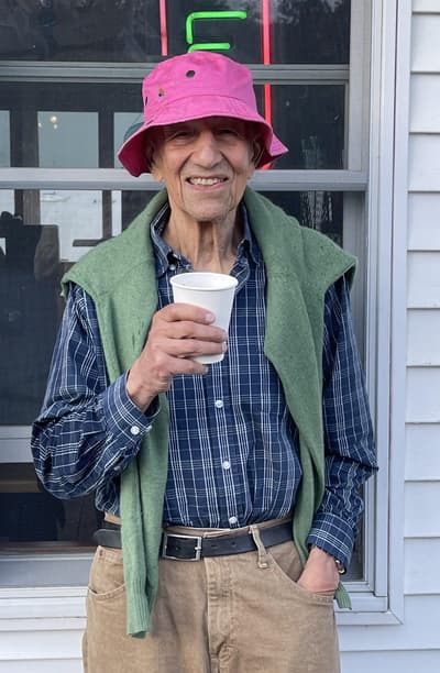 Photograph of Alex Katz smiling, holding a paper cup and wearing a pink bucket hat