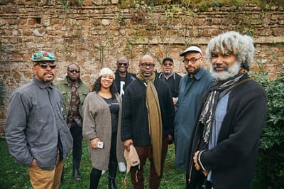 Eight artists standing together in front of a brick wall looking directly at the camera.