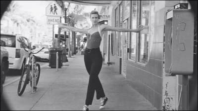 A still from a film of a dancer. A woman poses in the street with her legs elegantly crossed and arms stretched out.