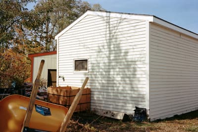 A color photograph of a yellow wheelbarrow in front of a white building surrounded by trees.