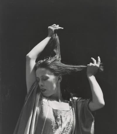 Black and white photograph of a woman holding her hair up in two separate parts, one clump in each hand. She looks down and to the rigght.