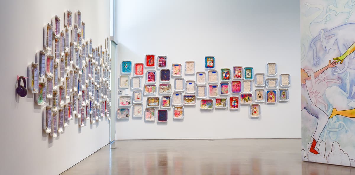 An art gallery with mixed media boxes and trays on the walls, and a painted abstract canvas visible on the right.