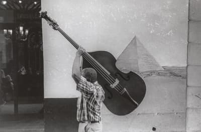Person holding an upright cello in front of a mural with a pyramid.