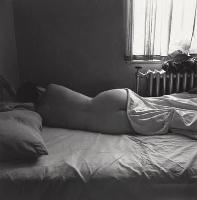 Black and white photograph of a nude woman lying in bed with her back facing towards the camera.