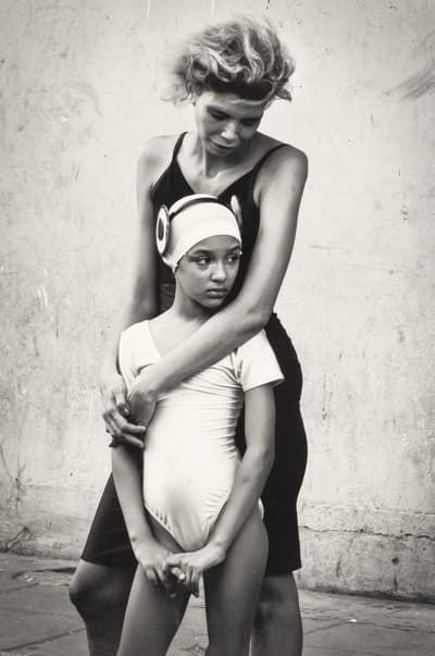 A person embraces a child in a leotard and swim cap against a textured wall.