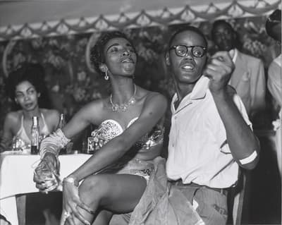 A lively nightclub scene with a woman and a man in conversation, surrounded by other patrons and elegant decor.