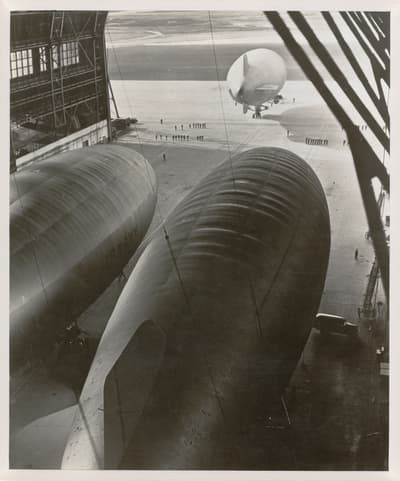 Two large airships inside a hangar with one labeled "U.S. Navy," and a third airship on the ground outside.