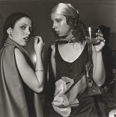 Two people in conversation at a social event, wearing headscarves and sleeveless garments.