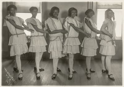 A vintage photograph of six young women in a line wearing matching outfits and holding each other's shoulders.
