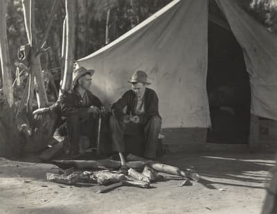 Two men sit in front of a canvas tent with a campfire setup in a wooded area.