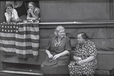 Four women in an urban setting, two leaning on a window sill with an American flag, two sitting below engaged in conversation.