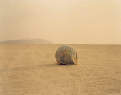 A large, covered sphere in a dry desert landscape with distant mountains and tire tracks.