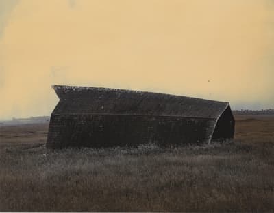 A weathered barn leaning in a grassy field under a muted yellow sky.