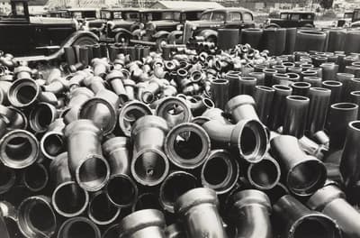 A black and white photo showing scattered metal pipes with vintage cars behind them.