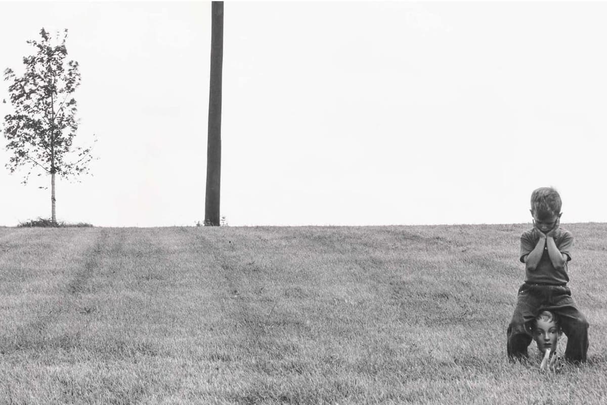 A young boy crouched on a grassy slope, resting his head on his crossed arms, with a mannequin head beneath him.