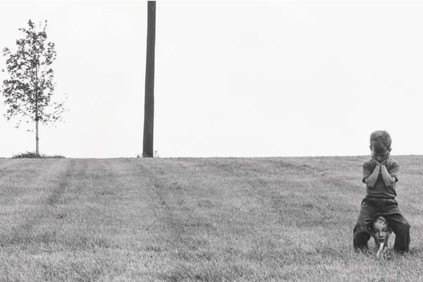 A young boy crouched on a grassy slope, resting his head on his crossed arms, with a mannequin head beneath him.