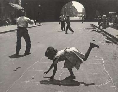 A young girl plays hopscotch on the street while others walk and play nearby.