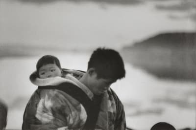 A black and white photo of a man carrying a baby on his back with a blurred natural background.