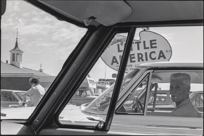 View through a car window showing a man in a neighboring car and a sign for “Little America.”