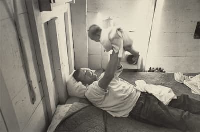 A man joyfully lifting a baby while lying on a bed in a simple, wooden-walled room.