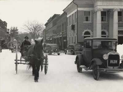 A snowy street scene with a horse-drawn cart and vintage cars alongside brick buildings.
