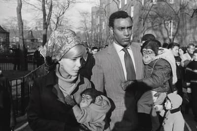 A man and a woman hold dressed chimpanzees in an urban setting with leafless trees and a crowd in the background.