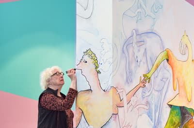 A photograph of Gladys Nilsson intently painting "Rounding Rosie's Ring: Dance, You Fools, Dance" directly on the wall in the gallery.