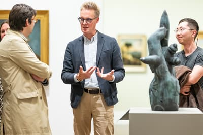 Three men conversing in a gallery next to a figurative sculpture.