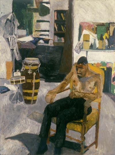 Portrait of a shirtless man sitting in a chair infant of two drums and shelves filled with books.